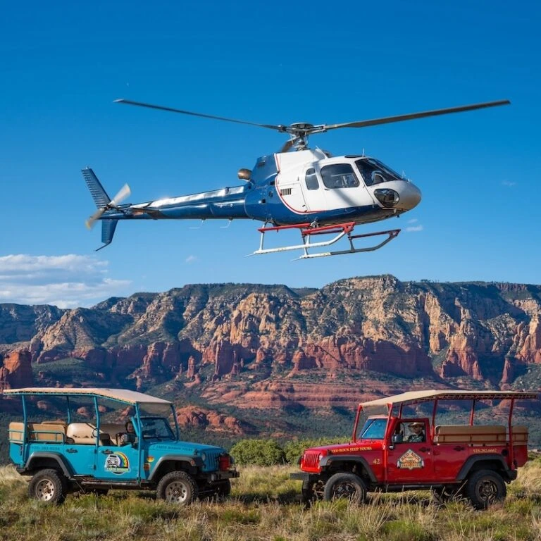 Helicopter flying above two off-road jeeps in a scenic desert landscape with Sedona red rock formations in the background.