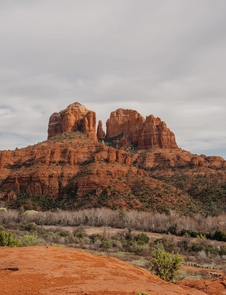 Red rock formation in Sedona, Arizona, surrounded by bushes and trees under a cloudy sky.