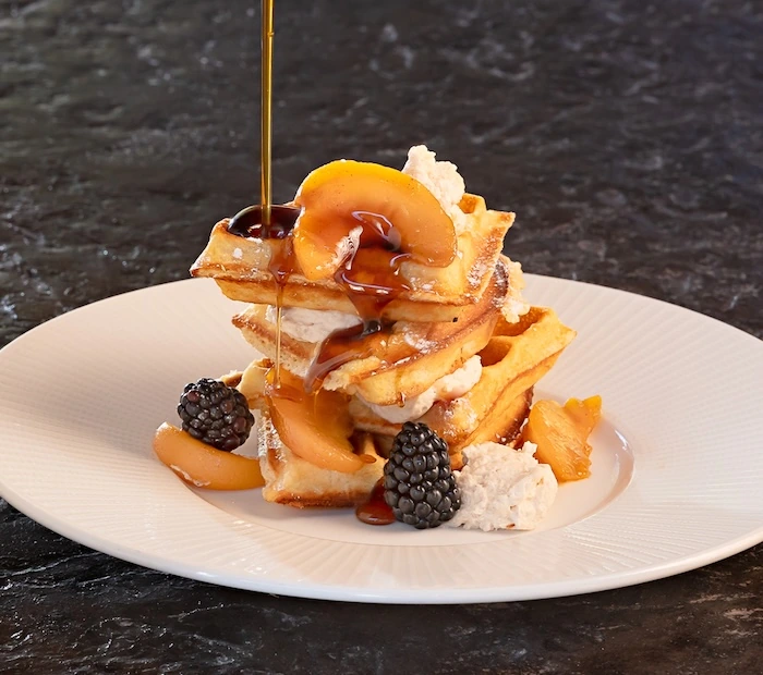 Stack of waffles topped with peach slices, blackberries, cream, and syrup being poured, served on a white plate on a dark surface.