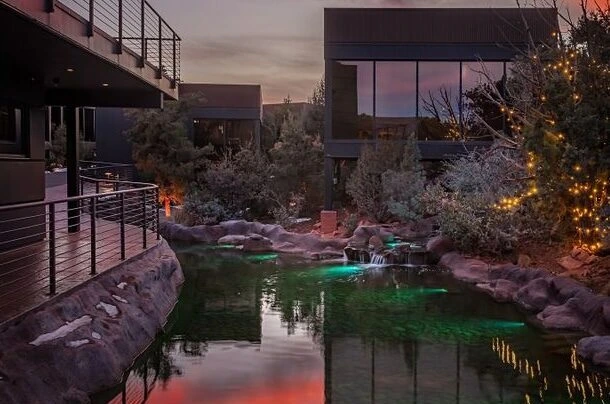 Outdoor pond with green lighting, surrounded by modern resort buildings, rocky edges, trees, and string lights at sunset.