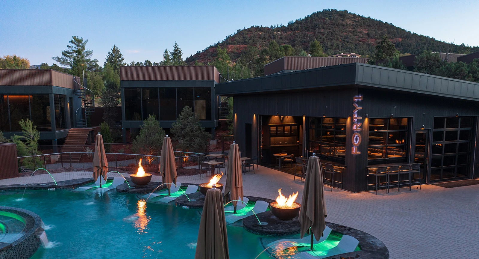 A pool area with fire pits and umbrellas at dusk, with a mountain view.
