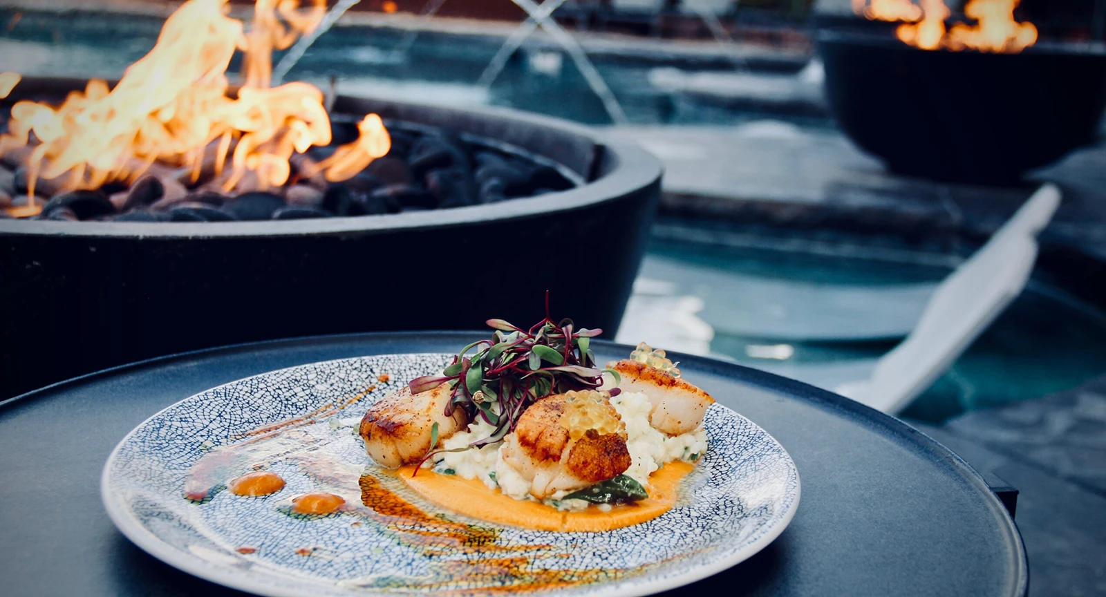 a plate of food with scallops, microgreens, and sauce in front of a fire pit