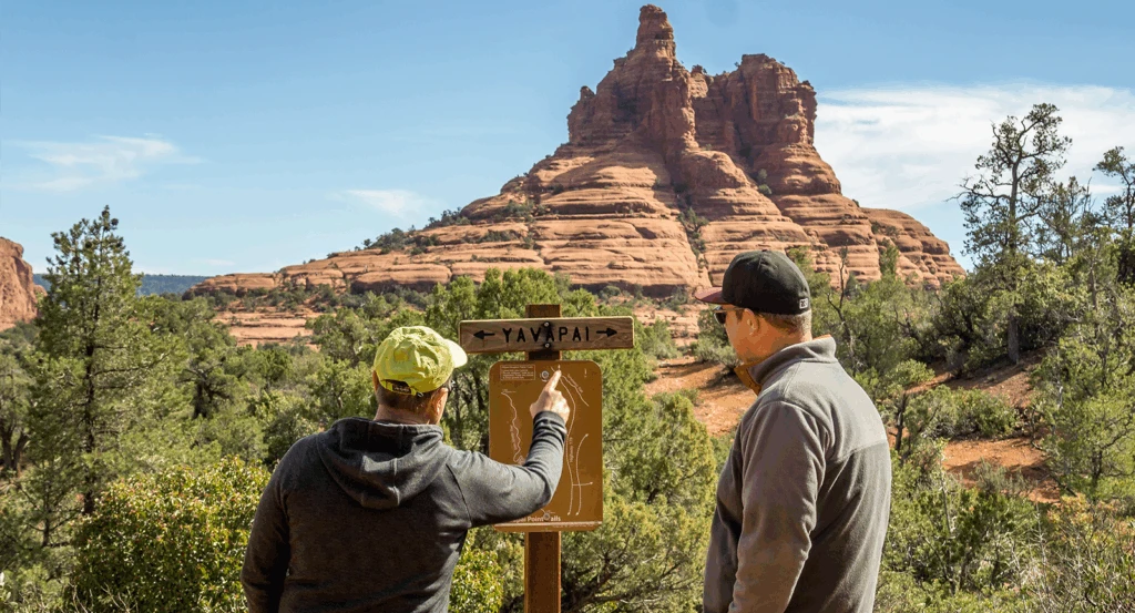 Two men on easy hikes in Sedona looking at a sign with red rocks in the background.