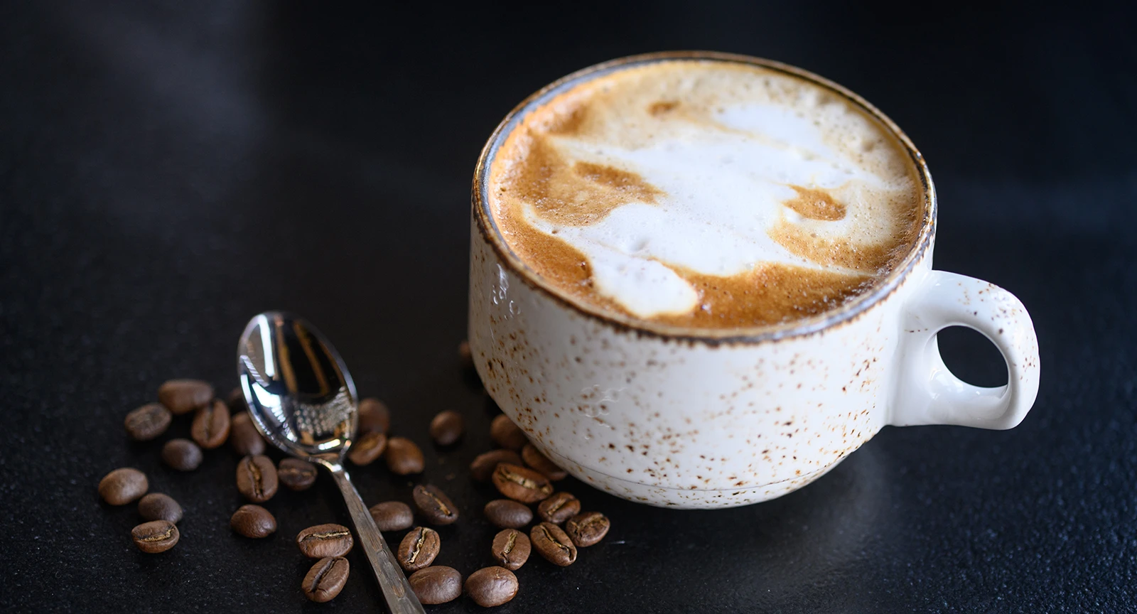 A cup of coffee with milk foam and coffee beans on a dark surface